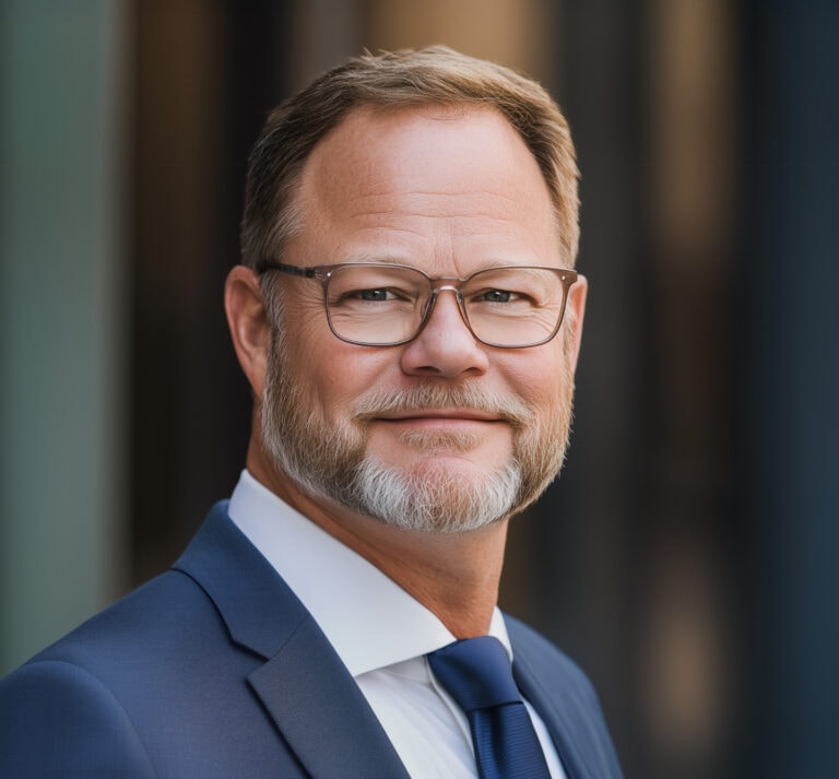 Professional portrait of a middle-aged man with glasses, wearing a navy blue suit and tie, smiling confidently outdoors.