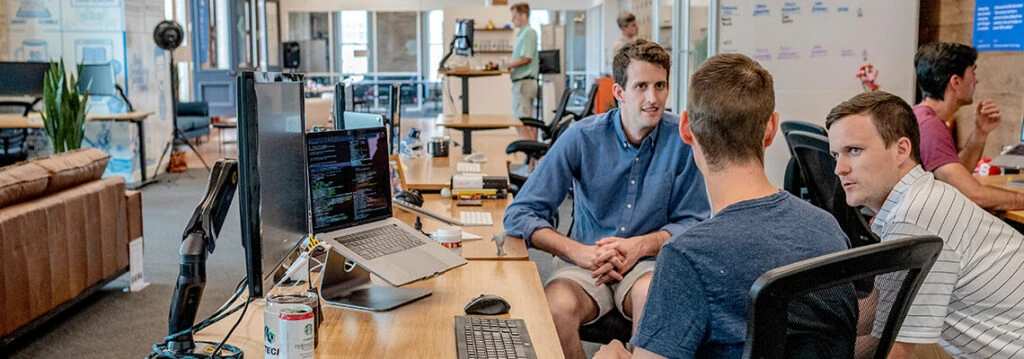 Three young men engaged in a discussion in a modern office, with computers and monitors on the desk, and a whiteboard in the background.